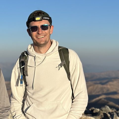 Joe Whittle smiling on a rocky mountain summit, wearing a headlamp and a daypack.